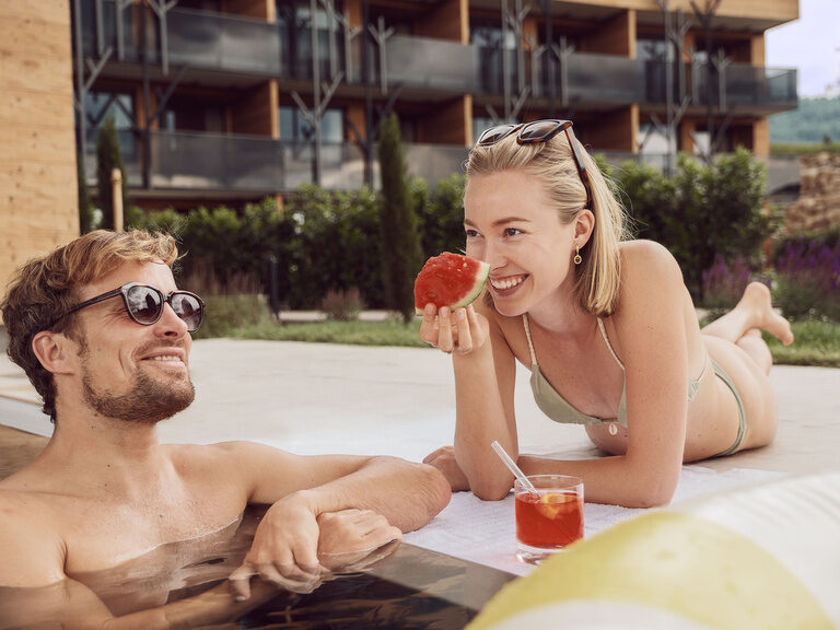Ein glückliches Paar entspannt am Außenpool des Tonihof und genießt köstliche Speisen und bunte Cocktails vor der stimmungsvollen Kulisse des Hotels.