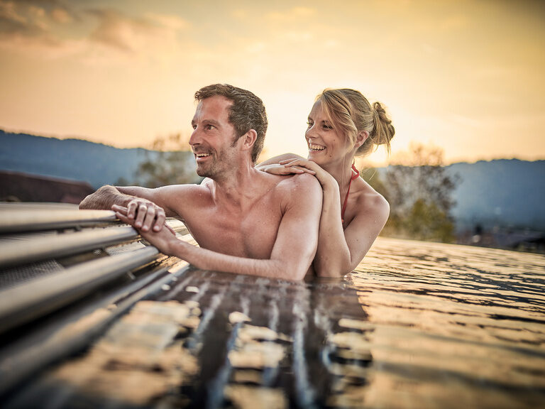 Ein glückliches Paar entspannt am Außenpool des Tonihof und genießt den beeindruckenden Panoramablick auf die umliegende Landschaft des Bayerischen Waldes.