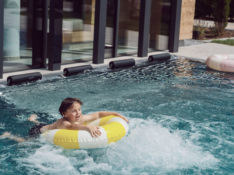 Ein Kind treibt fröhlich im Schwimmreifen über aufsteigende Blasen im klaren Wasser des Außenpools.